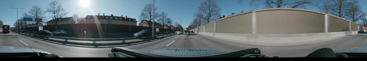 A clear spring day in Stockholm, Sweden, shows a car following traffic along a road lined with bare trees, red buildings, and grey sound walls.
