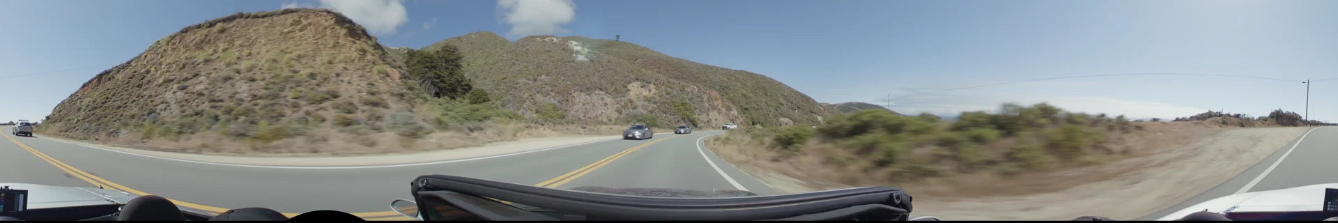 A winding road in Big Sur with oncoming traffic, bordered by steep, brush-covered hills under a clear afternoon sky.