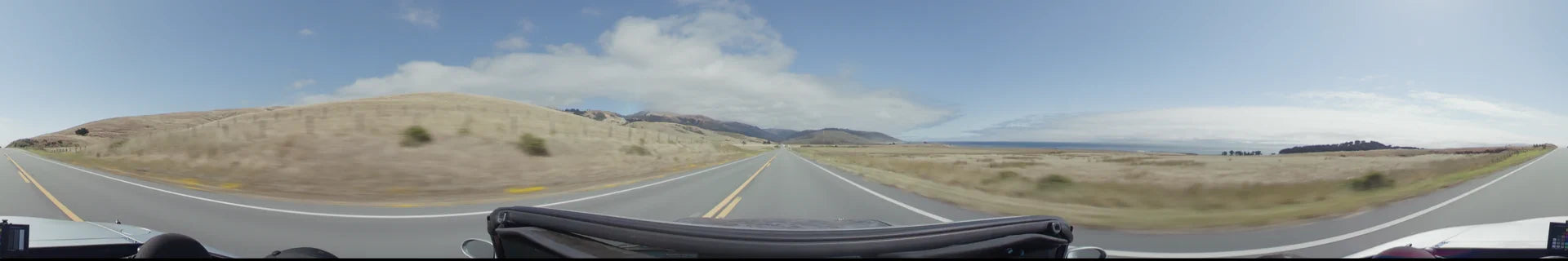 A straight two-lane highway in Big Sur stretches toward distant mountains, bordered by expansive golden grasslands and a bright, open sky.