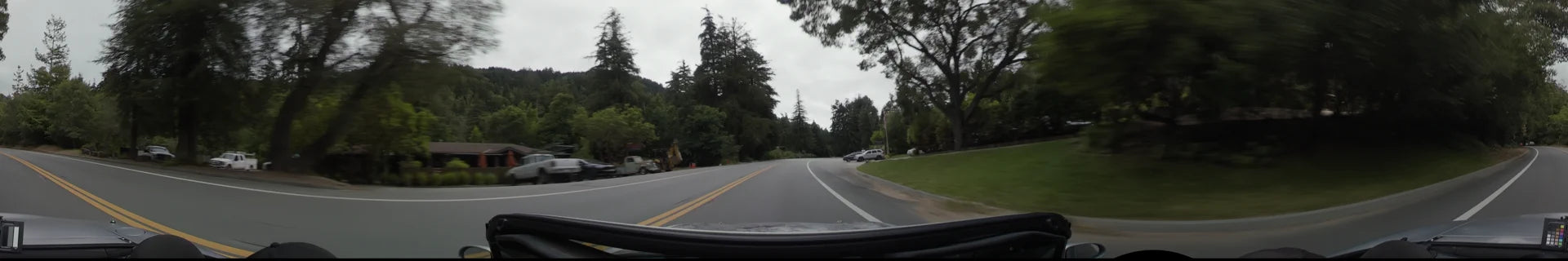 A winding road in Big Sur flanked by tall evergreen trees and lush greenery, with parked cars on a roadside shoulder and a construction vehicle nearby.