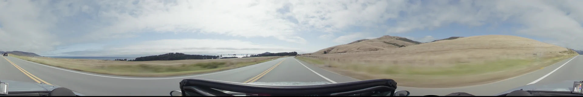 A two-lane highway in Big Sur stretches past golden grassy hills and a distant coastline under a clear, bright sky.