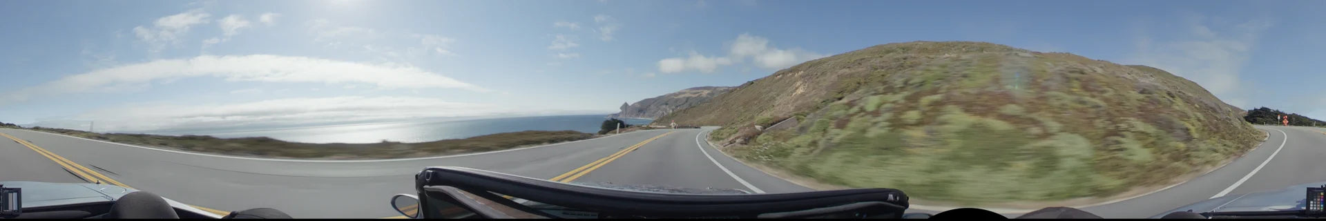 A winding coastal road in Big Sur curves along steep, grassy cliffs, offering a wide view of the blue ocean and distant mountains under a clear sky.