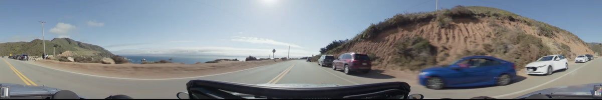 A two-lane highway in Big Sur with several cars parked along a steep, scrubby hillside, bordered by a wide shoulder and the distant coastline.