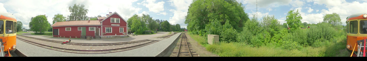 A daytime view from a train in Uppsala, Sweden, showing tracks, a concrete platform, and dense green summer foliage under a partly cloudy sky.