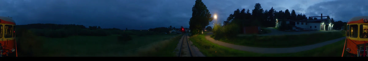 View from a train in Uppsala, Sweden, at partly cloudy summer dusk. Railway tracks lead past houses and green foliage with a glowing street lamp and signal.