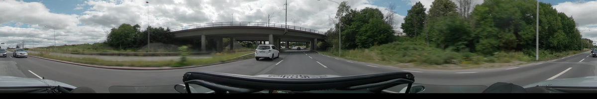 A concrete overpass spans a multi-lane road in Uppsala, with a white car driving ahead and lush green trees lining the roadside.