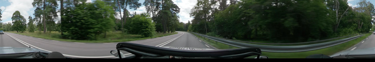 A curving road between Uppsala and Upplands-Bro is framed by dense green forests and a metal guardrail under a partly cloudy sky.
