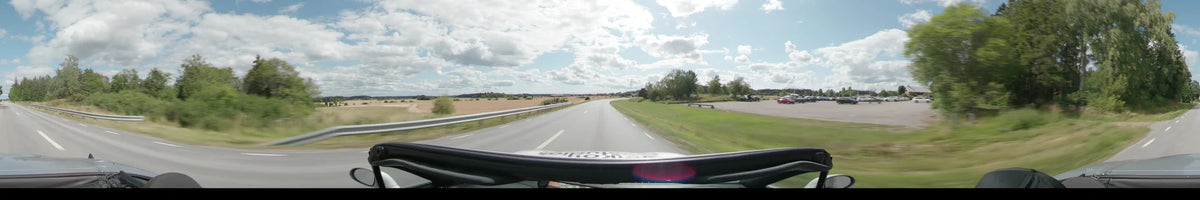 A multi-lane road between Uppsala and Upplands-Bro curves past golden fields, green grass, and scattered trees under a partly cloudy sky.