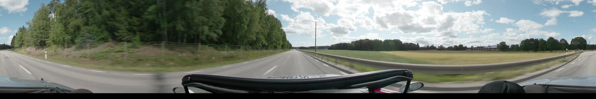 A straight road in Upplands-Bro is bordered by a metal guardrail, open green fields, and dense forests under a partly cloudy sky.