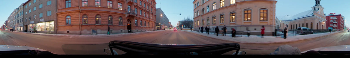 A car drives through a street in Uppsala, Sweden, on a clear winter afternoon. Colorful buildings line the road, with snow on the sidewalks and pedestrians nearby.