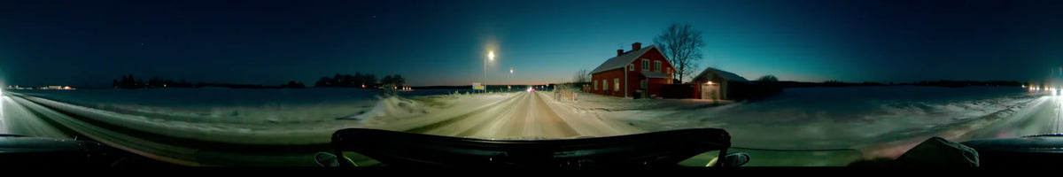 On a clear winter afternoon in Uppsala, Sweden, a car travels a snowy road past a red house and illuminated street lamps.