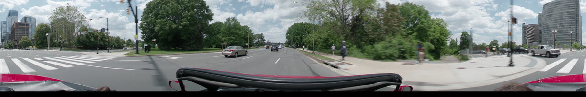 A car drives on a multi-lane road in Washington during a partly cloudy spring day, passing green trees and pedestrians with the Washington Monument in view.