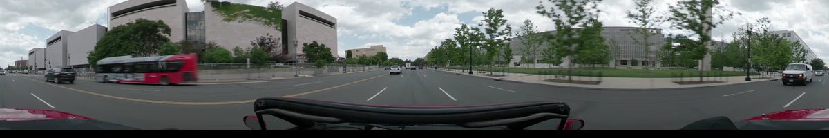 During a partly cloudy spring afternoon, a car drives past light-colored stone buildings and green trees on a wide road in Washington, United States.