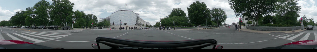 A large building under white scaffolding and green trees border a wide street in Washington, United States, as a car passes on a partly cloudy spring afternoon.