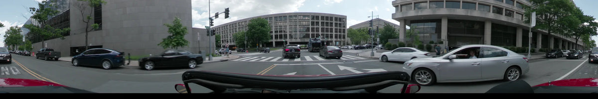 During a partly cloudy spring afternoon, a car drives through Washington, United States, passing a large grey building and lush trees at a wide intersection.
