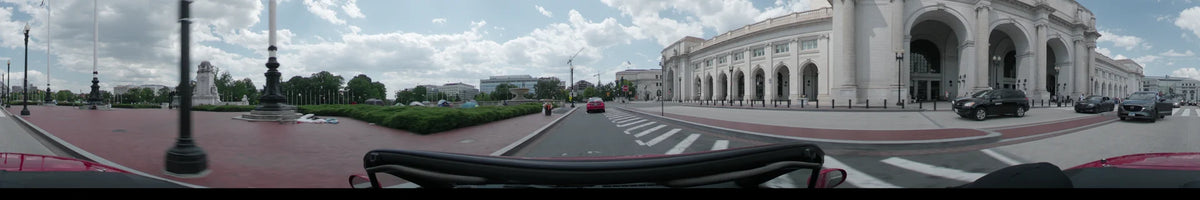 A car drives past the National Gallery of Art in Washington on a partly cloudy spring afternoon, featuring a city street with a crosswalk and greenery.