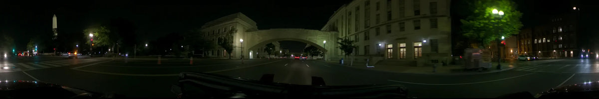 A wide stone archway between two massive illuminated buildings spans the road as a car travels through Washington, United States, on a clear spring night with distant traffic.