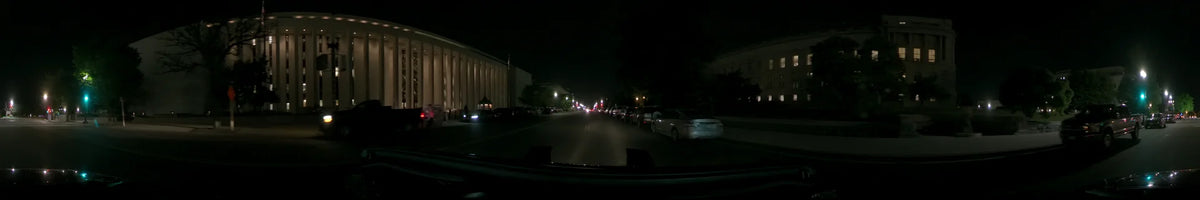 Parked cars and a large, white columnar building line the street as a car drives through Washington, United States, on a clear spring night.
