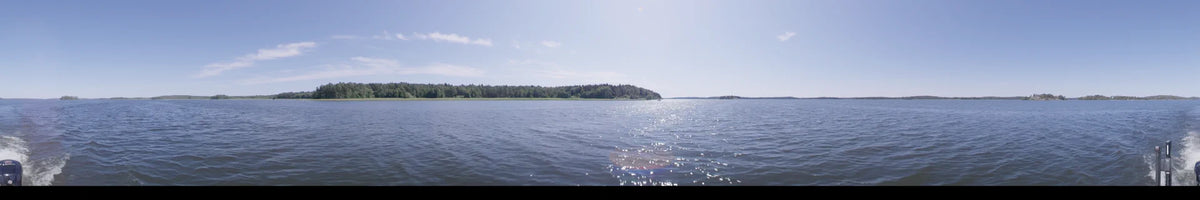 Sunlight sparkles across the water from a boat in Upplands-Bro, Sweden. A dense green forest borders the lake under a clear summer sky during the day.