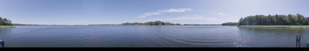 The blue waters of a lake in Upplands-Bro, Sweden, meet a distant wooded shoreline. A clear summer sky overhead defines this daytime boat perspective.