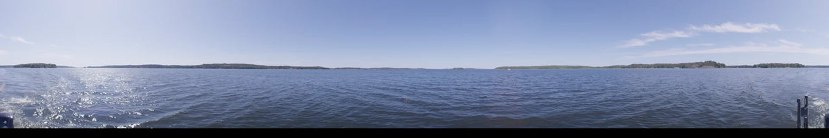 Blue water and a far-off green treeline define this daytime boat perspective in Upplands-Bro, Sweden, beneath a clear summer sky.