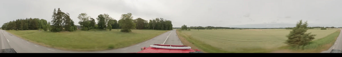 On an overcast summer morning in Märsta, Sweden, a bus drives past lush green fields and wooded areas along a paved two-lane road.