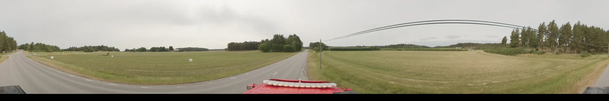 In Märsta, Sweden, a bus travels along a straight road on an overcast summer morning, passing through a landscape of wide green fields and forests.