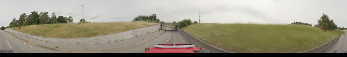 Concrete walls and grassy slopes flank the road as a red bus passes under a bridge in Märsta, Sweden, on an overcast summer morning.