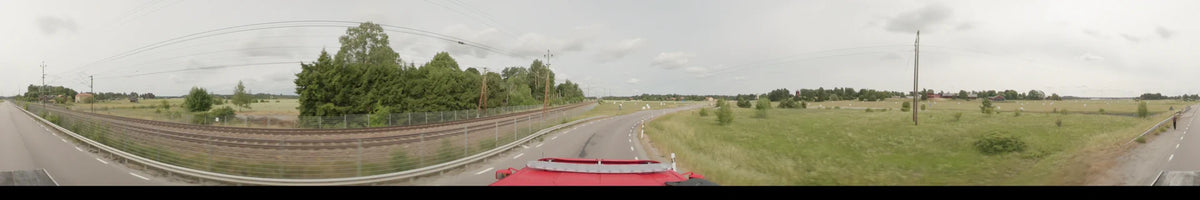 Railway tracks run parallel to a curved road as a red bus travels through the green landscape of Märsta, Sweden, on an overcast summer morning.