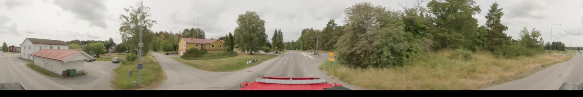Between dense trees and a bus stop, a road stretches forward for a red bus in Märsta, Sweden, on an overcast summer morning.