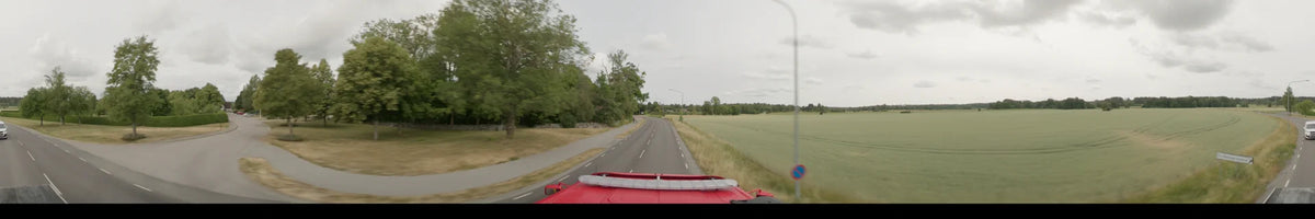 A bus travels a road in Märsta, Sweden, on an overcast summer morning, with thick trees on the left and open fields on the right.