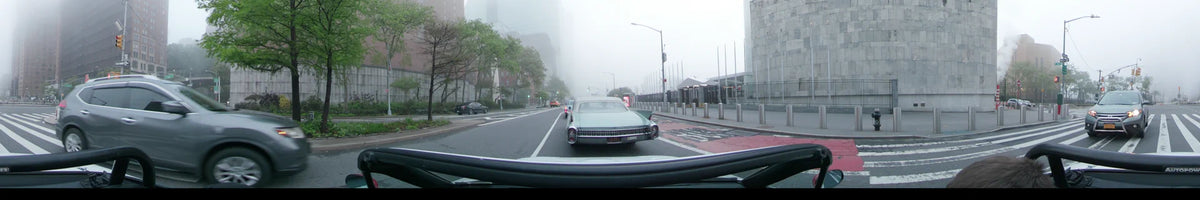 Dense fog blankets a New York street on a spring morning. A vintage car drives forward, passing budding trees and city infrastructure in the United States.