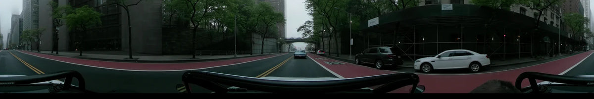 Green spring trees line a New York street on an overcast morning. A car travels behind a vintage blue vehicle toward a concrete bridge overpass.