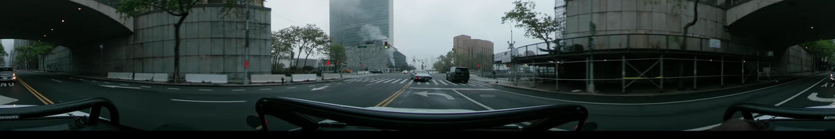 One World Trade Center looms over a wide street in New York during an overcast spring morning as a car drives past traffic and modern architecture.