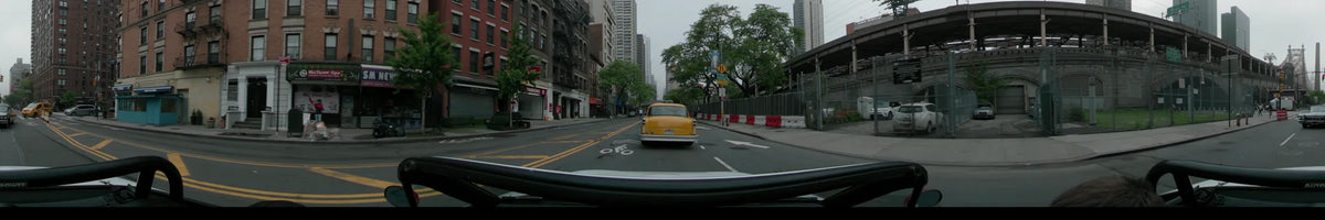 Following a yellow car through New York on an overcast spring morning, the street is lined with brick buildings, green trees, and red and white barriers.