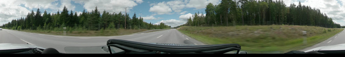 A multi-lane road in Upplands-Bro is flanked by dense green forests and grassy embankments under a partly cloudy sky.
