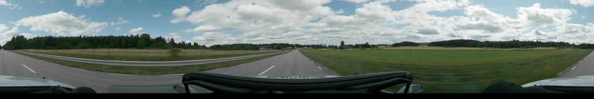 A multi-lane road between Upplands-Bro and Uppsala is flanked by green fields, fences, and distant red farm buildings under a partly cloudy sky.
