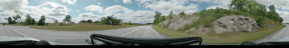 A paved road curves between open fields and a rocky hillside with green trees under a partly cloudy sky between Upplands-Bro and Uppsala.