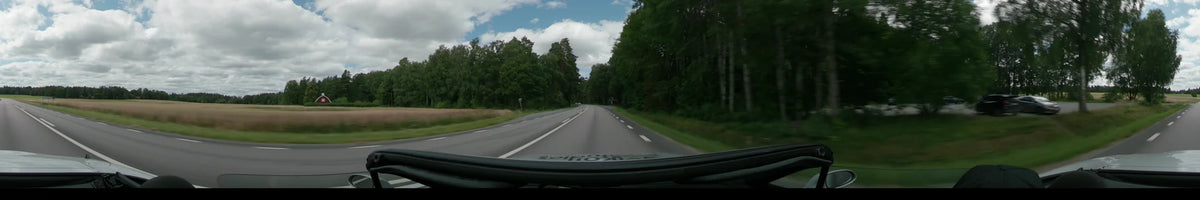 A multi-lane road winds through a dense green forest under a partly cloudy sky, with a white car visible in the distance.