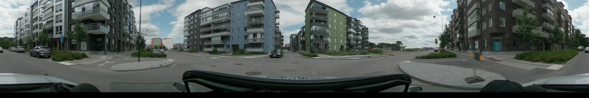 Modern blue and green residential buildings line a street in Uppsala, with a dark car driving ahead on the asphalt road under a partly cloudy sky.