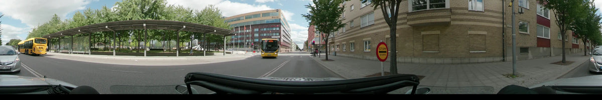 A yellow bus drives along a city street in Uppsala, flanked by a large modern office building and brick residential architecture with leafy green trees.
