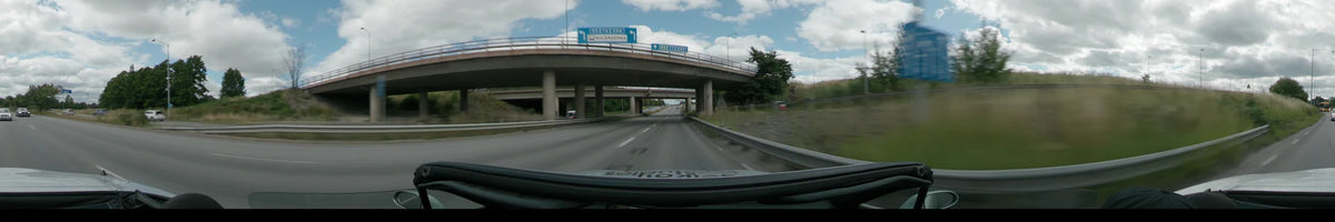 A multi-lane highway in Uppsala passes under a concrete overpass with large blue overhead traffic signs, bordered by green foliage and a metal guardrail.