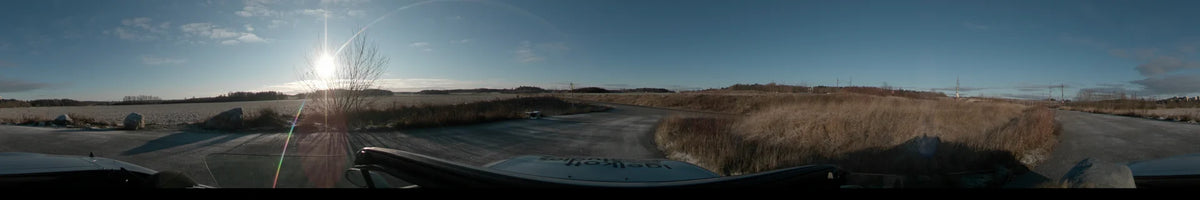 A car travels on a paved road through the rural countryside of Lennartsnäs, Sweden, during a clear autumn day, surrounded by flat fields and dry grass.