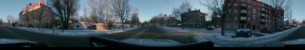 A clear winter dawn illuminates a residential road in Stockholm, Sweden, as a car passes snow-covered ground, bare trees, and apartment buildings.