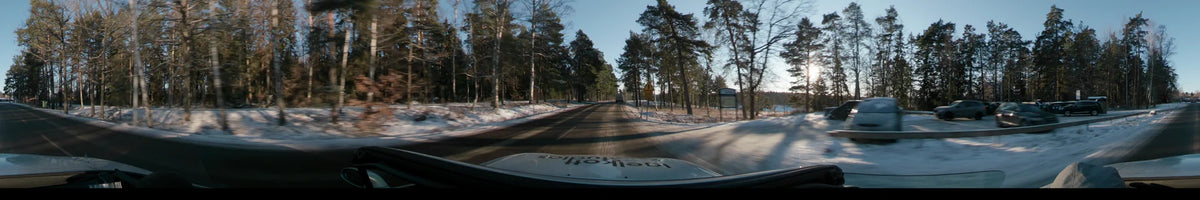 A clear winter dawn fills the sky in Stockholm, Sweden, as a car follows a truck past tall, snow-covered pine trees.