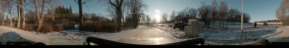 A car drives on a snowy road in Stockholm, Sweden, during a clear winter sunset, with bare trees lining the path under a bright sky.