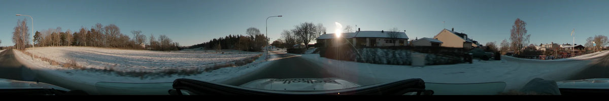A car drives along a residential road in Stockholm, Sweden, during a clear winter sunset, with snow on the ground and houses under a bright sky.