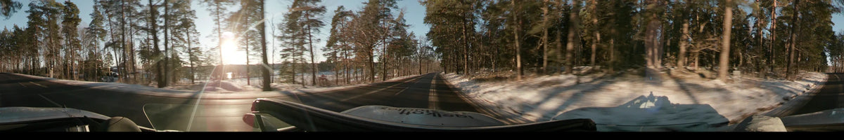 Through the forests of Stockholm, Sweden, a car travels along a road with snow-covered shoulders under a clear sky during a winter sunset.