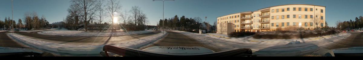 Residential houses and bare trees line a snow-covered street in Stockholm, Sweden, during a clear winter sunset as seen from a car.