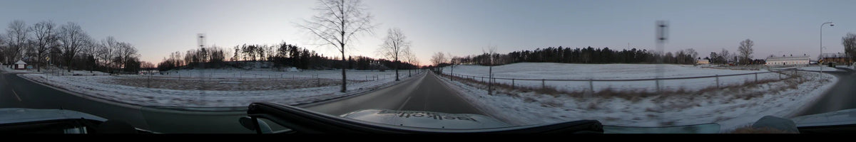 Bare trees and snowy fields line a rural road in Stockholm, Sweden, where a car drives under a clear winter sunset.